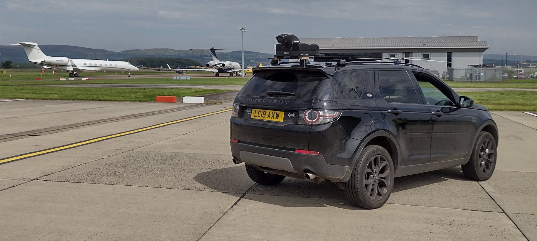 An Atlas survey vehicle with a roof-mounted sensor rig on an airport apron, aircraft parked in the background.